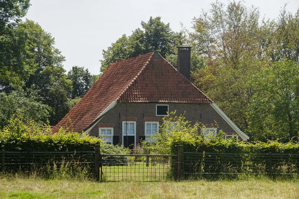 Bakstenen boerderij met rood pannendak achter een hekwerk en bomen