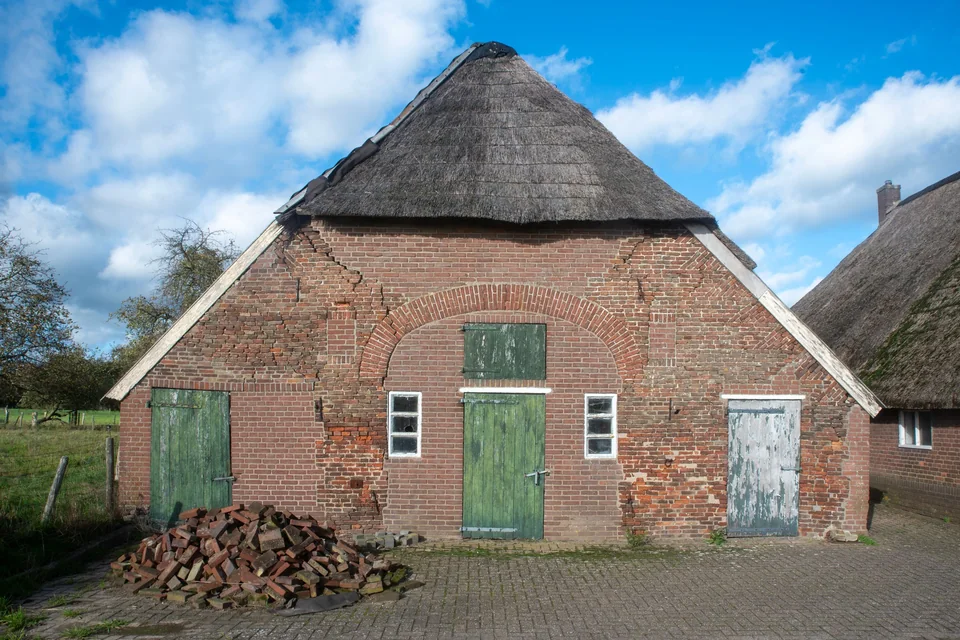Bakstenen schuur met rieten kap en groene deuren op het erf van een boerderij